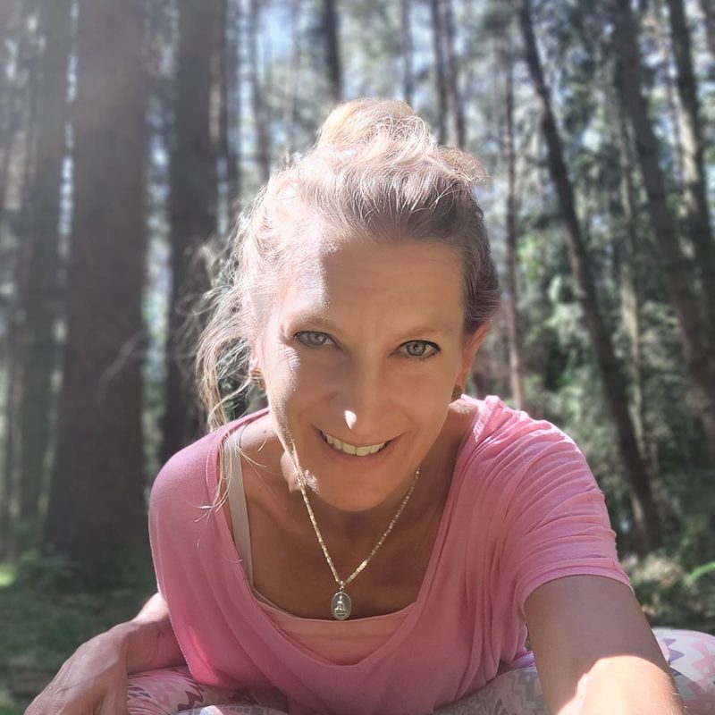 Alice sitzt lächelnd auf moosigem Waldboden in Yoga-Kleidung, hinter ihr hohe Bäume